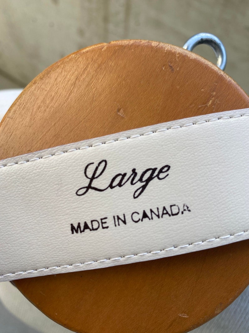 May include: Close-up of a white leather belt with the word "Large" in cursive script. The text "MADE IN CANADA" is printed below. The belt is displayed on a brown wooden form.