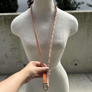 May include: A thin orange leather belt with a silver buckle. The belt is on a white mannequin.
