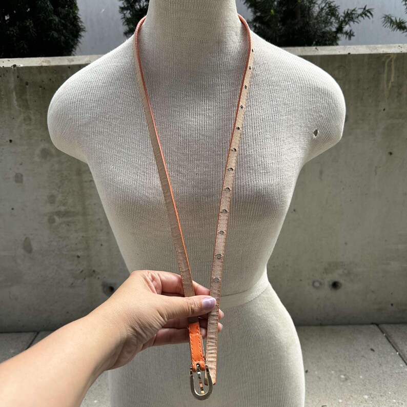 May include: A thin orange leather belt with a silver buckle. The belt is on a white mannequin.