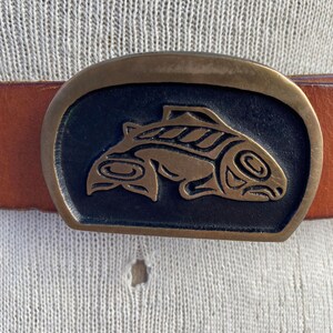 May include: A brown leather belt with a brass buckle featuring a stylized salmon design.