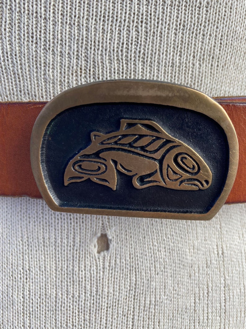 May include: A brown leather belt with a brass buckle featuring a stylized salmon design.