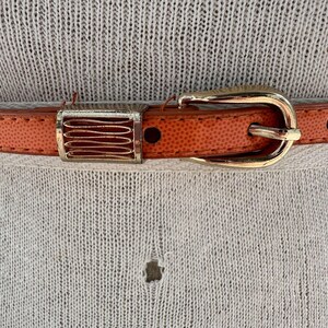 May include: A close-up of a brown leather belt with a gold buckle. The belt has a textured surface and a decorative metal piece.