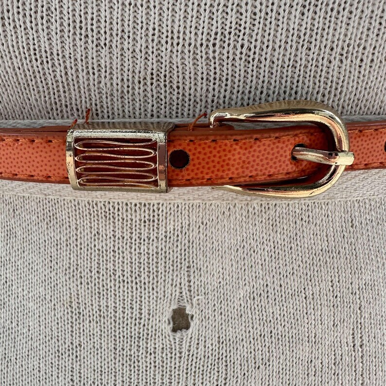 May include: A close-up of a brown leather belt with a gold buckle. The belt has a textured surface and a decorative metal piece.
