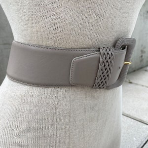 Puede incluir: Un cintur&oacute;n ancho de cuero gris claro con una hebilla trenzada. La hebilla es de color gris claro y tiene un acento de metal dorado.