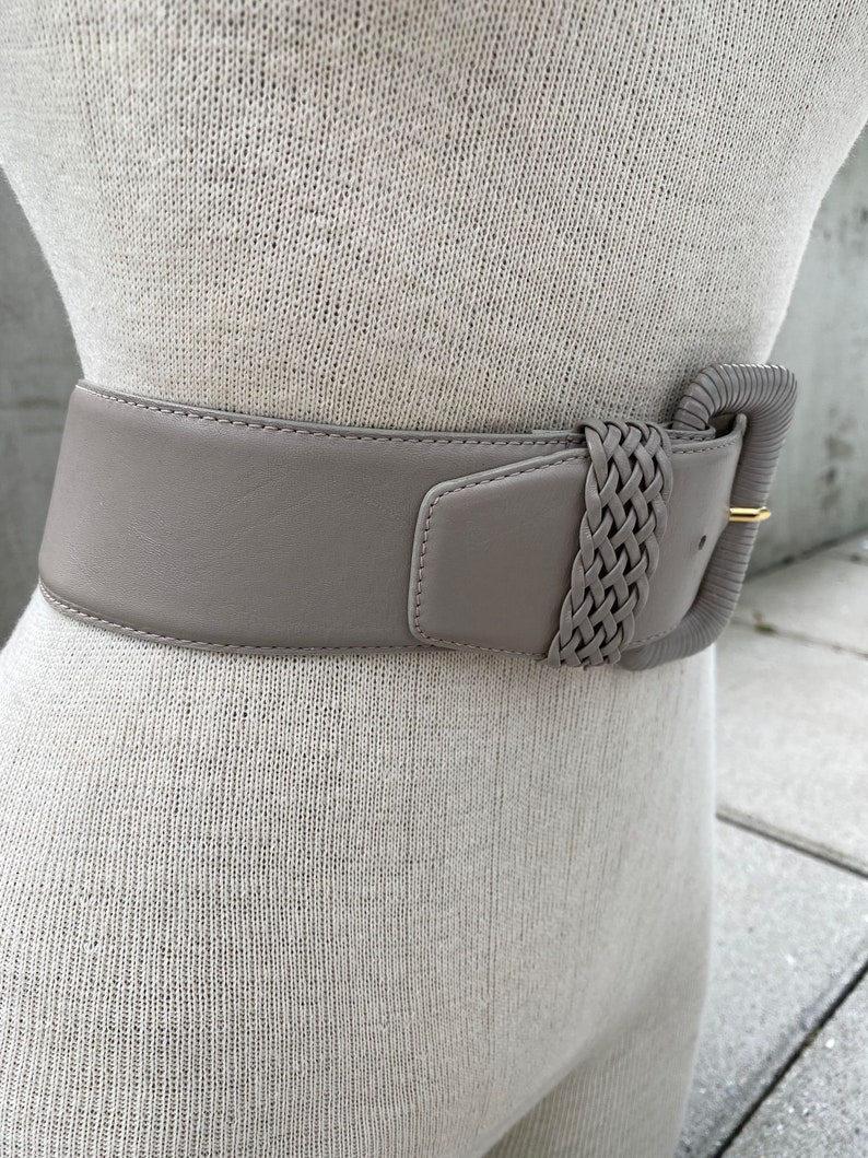 Puede incluir: Un cintur&oacute;n ancho de cuero gris claro con una hebilla trenzada. La hebilla es de color gris claro y tiene un acento de metal dorado.