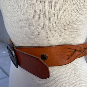 May include: A brown leather belt with a braided design and a silver buckle. The buckle has a decorative design.