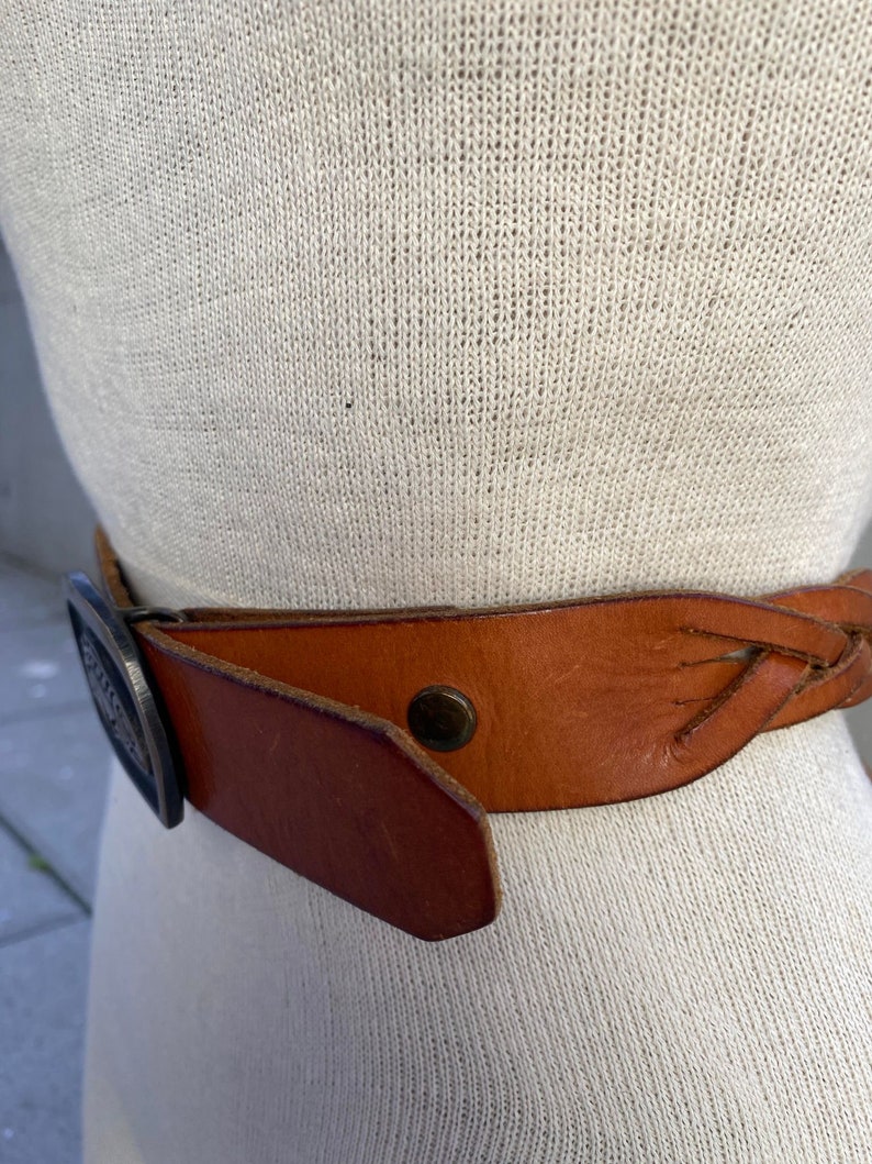 May include: A brown leather belt with a braided design and a silver buckle. The buckle has a decorative design.