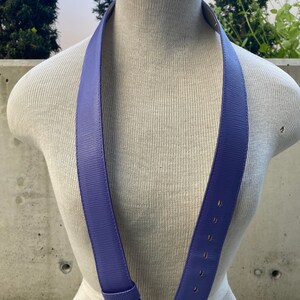 May include: A periwinkle blue belt with a rectangular buckle and a series of evenly spaced holes. The belt is displayed on a mannequin, showcasing its design and texture. The buckle is silver.