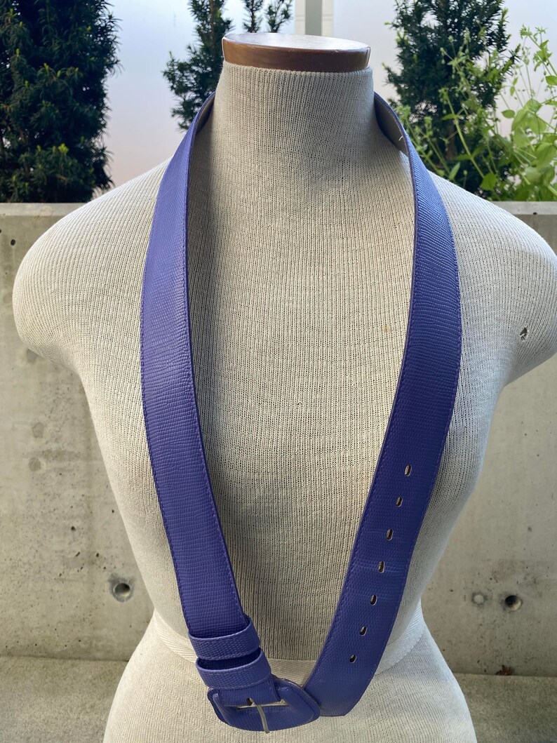 May include: A periwinkle blue belt with a rectangular buckle and a series of evenly spaced holes. The belt is displayed on a mannequin, showcasing its design and texture. The buckle is silver.