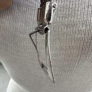 Peut inclure: Un pendentif en m&eacute;tal argent&eacute; de forme triangulaire, attach&eacute; &agrave; un fermoir en m&eacute;tal. L'arri&egrave;re-plan est un tissu textur&eacute; gris clair.