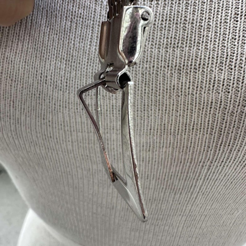Peut inclure: Un pendentif en m&eacute;tal argent&eacute; de forme triangulaire, attach&eacute; &agrave; un fermoir en m&eacute;tal. L'arri&egrave;re-plan est un tissu textur&eacute; gris clair.