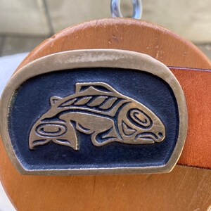 May include: A brass belt buckle with a stylized fish design in black and gold. The buckle is attached to a brown leather belt.