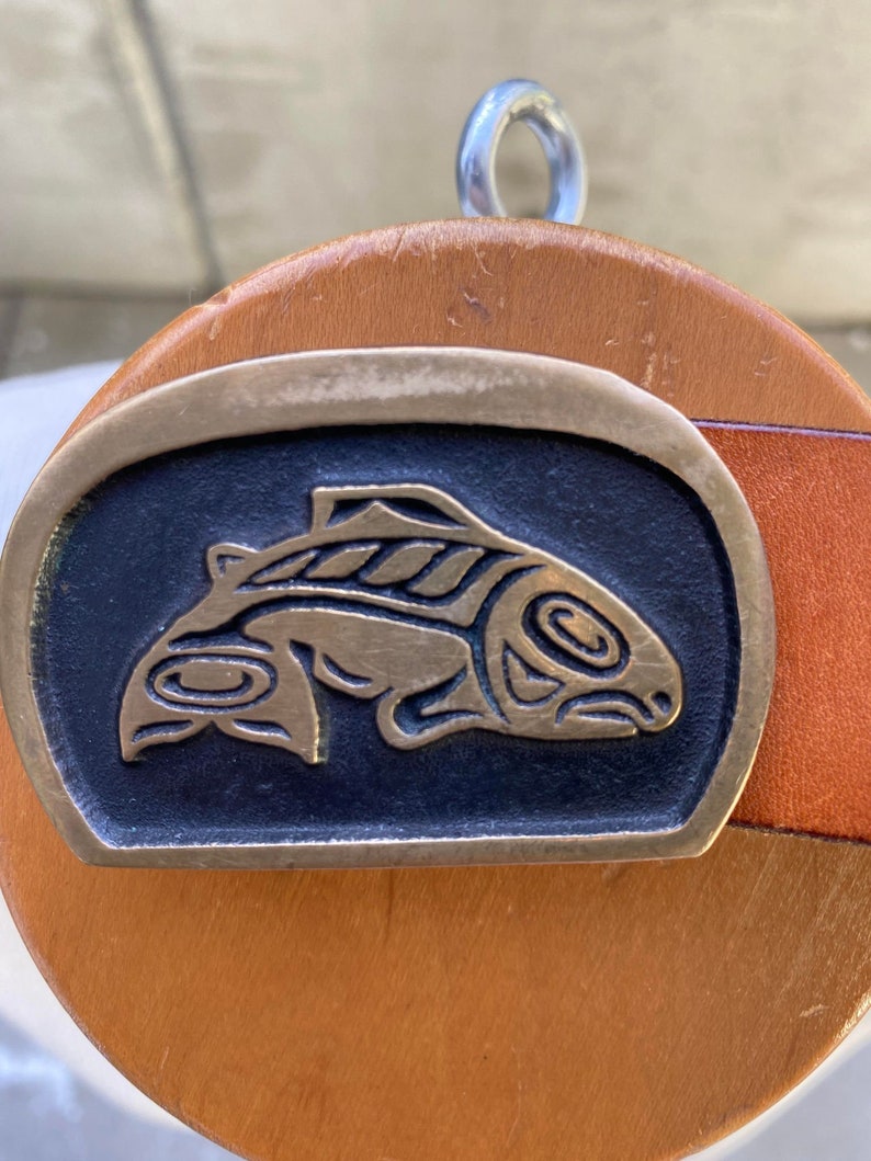 May include: A brass belt buckle with a stylized fish design in black and gold. The buckle is attached to a brown leather belt.