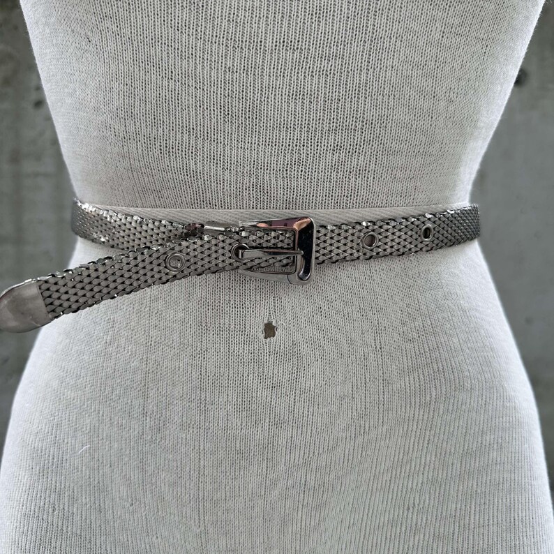 Peut inclure: Une ceinture en maille argent&eacute;e avec une boucle rectangulaire et une extr&eacute;mit&eacute; pointue. La ceinture est enroul&eacute;e autour d'un mannequin, mettant en valeur son design et son ajustement. La ceinture comporte une s&eacute;rie de petits trous pour le r&eacute;glage.