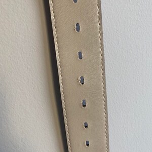 May include: A beige leather belt featuring a series of evenly spaced, vertically oriented oval adjustment holes. The belt is constructed with visible tonal stitching along both edges, running the full length of the visible strap section.