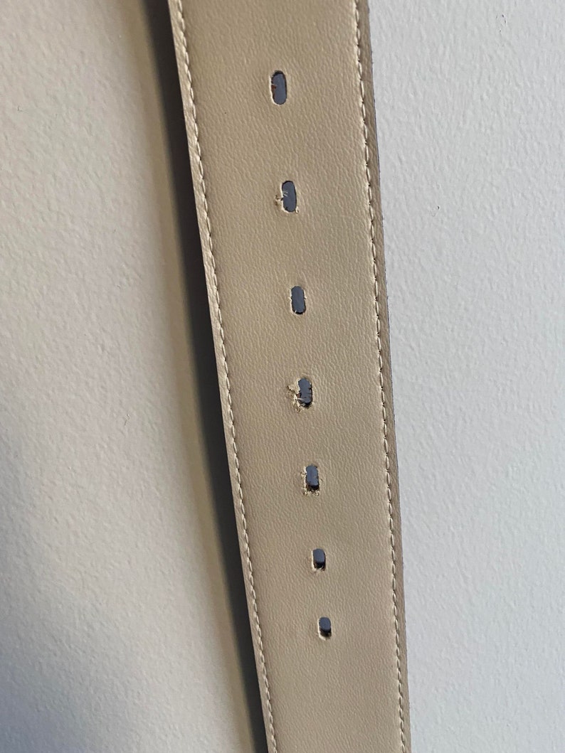 May include: A beige leather belt featuring a series of evenly spaced, vertically oriented oval adjustment holes. The belt is constructed with visible tonal stitching along both edges, running the full length of the visible strap section.