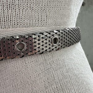 Peut inclure: Une ceinture en m&eacute;tal argent&eacute; avec un motif en maille. La ceinture comporte deux &oelig;illets et une s&eacute;rie de pi&egrave;ces imbriqu&eacute;es en forme d'&eacute;cailles. La ceinture est pr&eacute;sent&eacute;e sur un tissu clair et textur&eacute;.