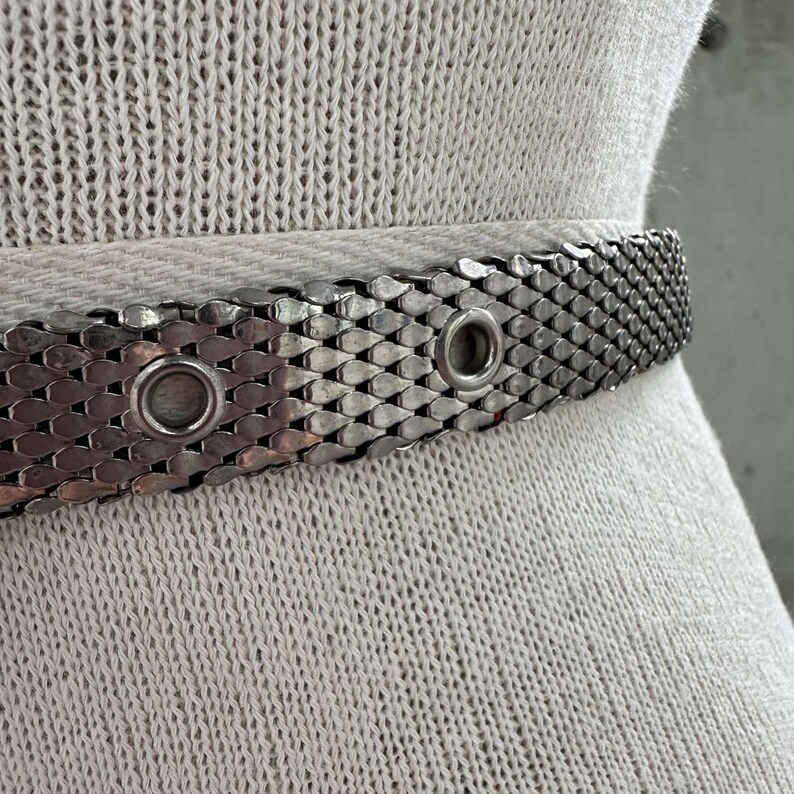 Peut inclure: Une ceinture en m&eacute;tal argent&eacute; avec un motif en maille. La ceinture comporte deux &oelig;illets et une s&eacute;rie de pi&egrave;ces imbriqu&eacute;es en forme d'&eacute;cailles. La ceinture est pr&eacute;sent&eacute;e sur un tissu clair et textur&eacute;.