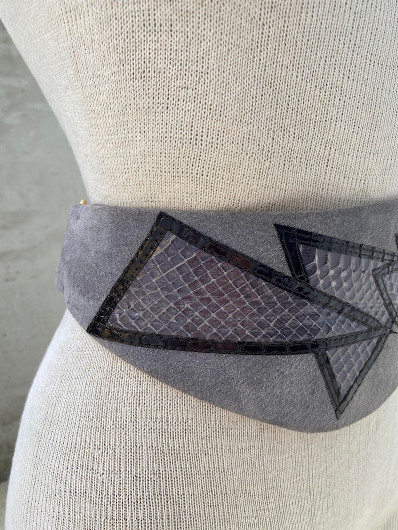 May include: A gray suede belt with black leather accents and a geometric design. The belt features a snakeskin pattern in the center.