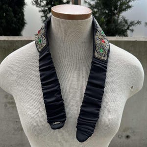 Pu&ograve; includere: Un girocollo nero con perline argentate e verdi. Il girocollo presenta un design decorativo con pietre rosse e verdi.