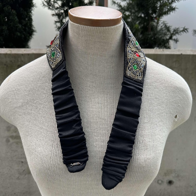 Pu&ograve; includere: Un girocollo nero con perline argentate e verdi. Il girocollo presenta un design decorativo con pietre rosse e verdi.