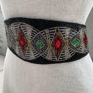 Pu&ograve; includere: Una cintura nera di perline con un design geometrico con accenti rossi, verdi e dorati. La cintura &egrave; adornata con grandi pietre preziose sfaccettate.