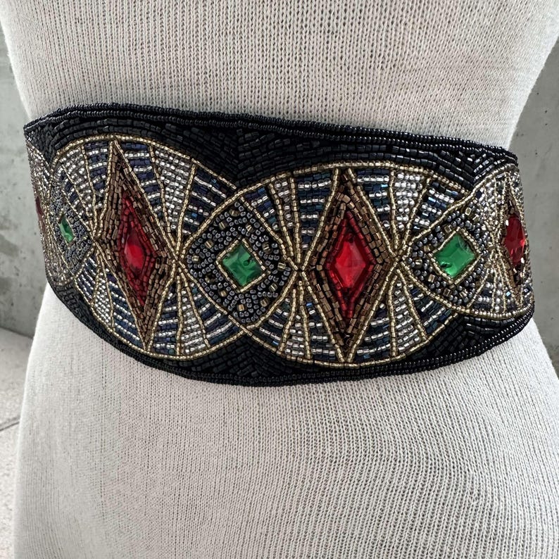 Pu&ograve; includere: Una cintura nera di perline con un design geometrico con accenti rossi, verdi e dorati. La cintura &egrave; adornata con grandi pietre preziose sfaccettate.