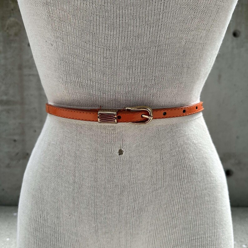 May include: A thin orange leather belt with a gold buckle. The buckle has a rectangular design with a raised pattern.
