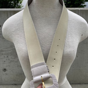 Puede incluir: Un cintur&oacute;n ancho de cuero blanco roto con una hebilla grande, rectangular y gris. La hebilla tiene un cierre de metal dorado.