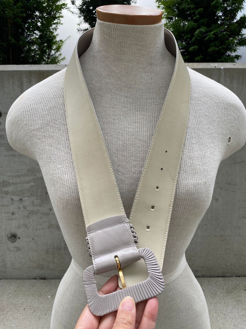 Puede incluir: Un cintur&oacute;n ancho de cuero blanco roto con una hebilla grande, rectangular y gris. La hebilla tiene un cierre de metal dorado.