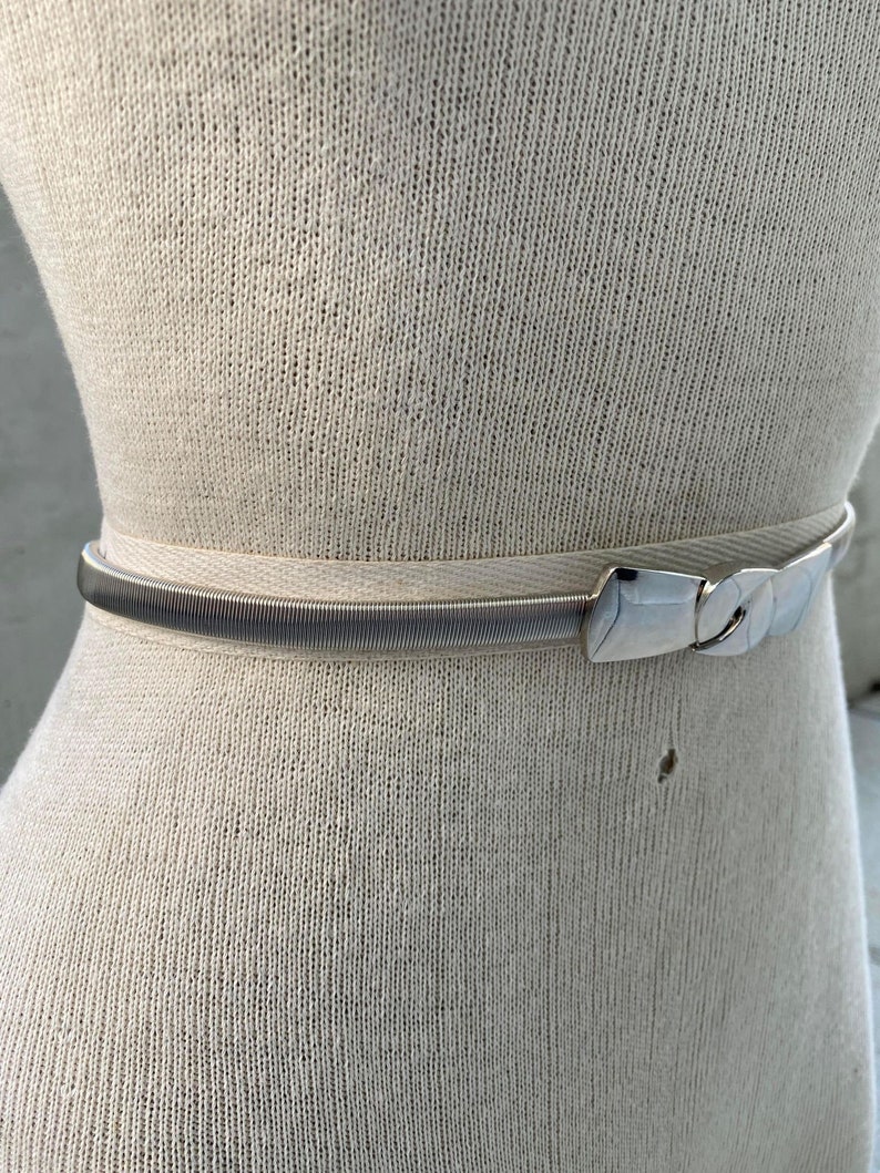 May include: A silver stretchy belt with a silver buckle. The belt is on a white mannequin.