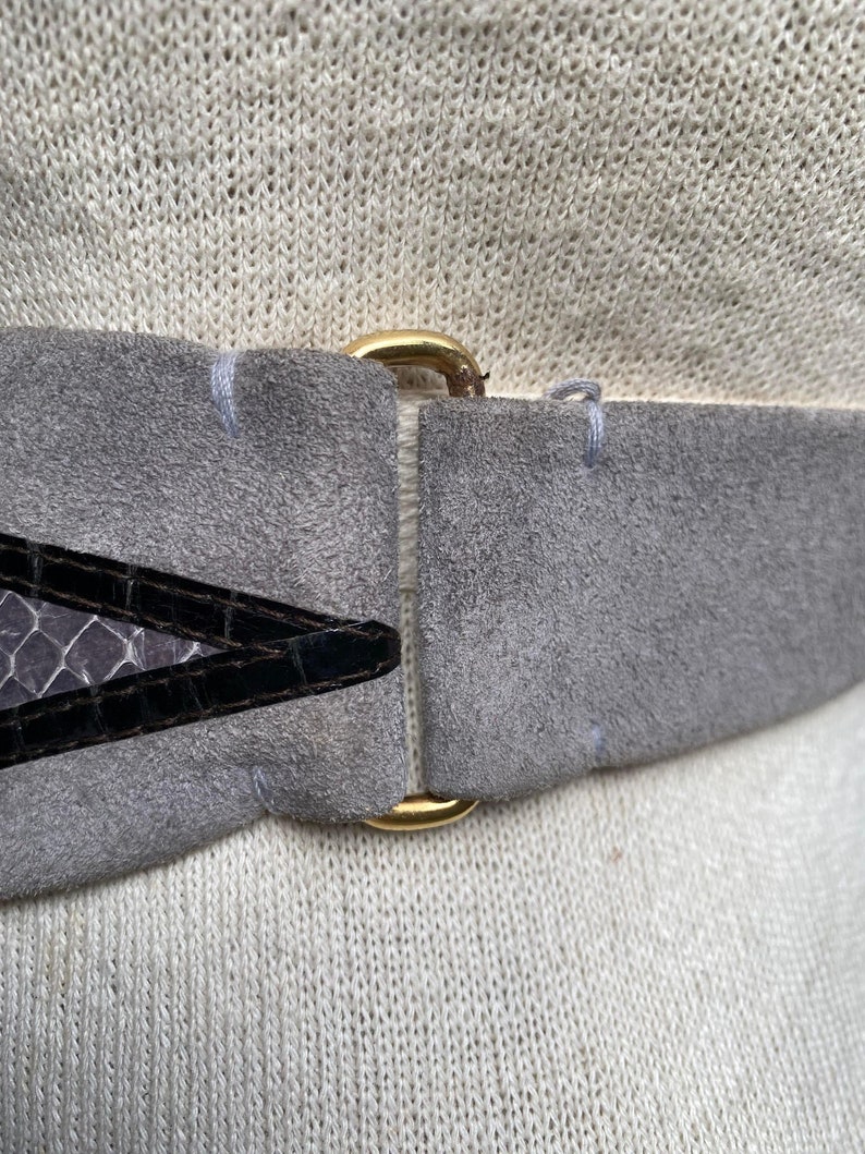 May include: A gray suede belt with a gold metal buckle and a black and gray snakeskin pattern on the front.