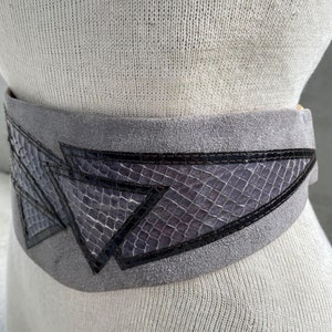 May include: A gray suede belt with a geometric design made of black and purple snakeskin. The belt is wide and has a buckle on the side.