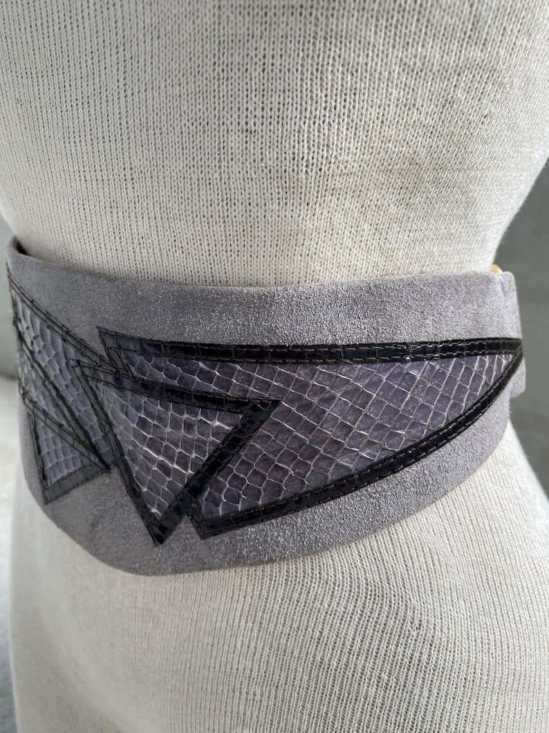 May include: A gray suede belt with a geometric design made of black and purple snakeskin. The belt is wide and has a buckle on the side.