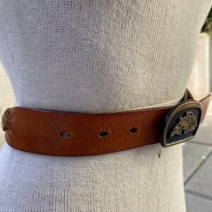 May include: A brown leather belt with a metal buckle featuring a stylized design. The buckle is gold-toned with black accents.