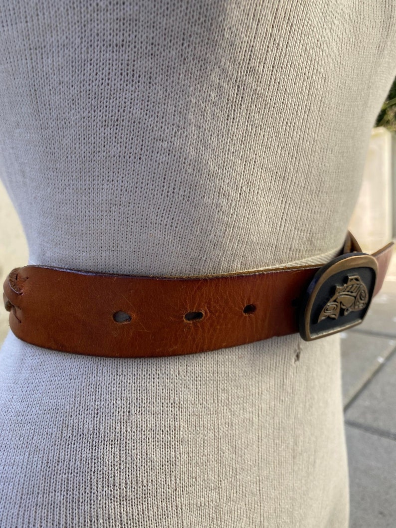May include: A brown leather belt with a metal buckle featuring a stylized design. The buckle is gold-toned with black accents.