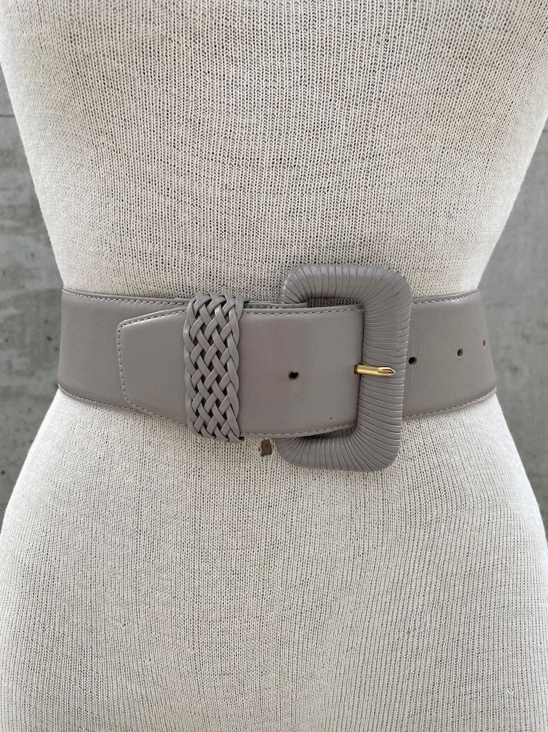 Puede incluir: Un cintur&oacute;n ancho de cuero gris con una hebilla grande y cuadrada. La hebilla tiene un dise&ntilde;o trenzado y un acabado dorado.