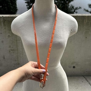 May include: A bright orange leather belt with a gold buckle. The belt is hanging on a white mannequin.