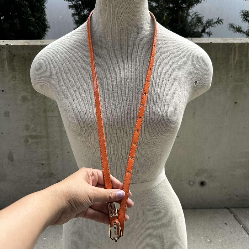 May include: A bright orange leather belt with a gold buckle. The belt is hanging on a white mannequin.