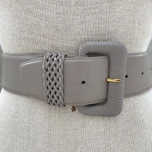 Puede incluir: Un cintur&oacute;n ancho de cuero gris con un detalle trenzado y una hebilla rectangular grande de color gris. La hebilla tiene un pasador de metal dorado.