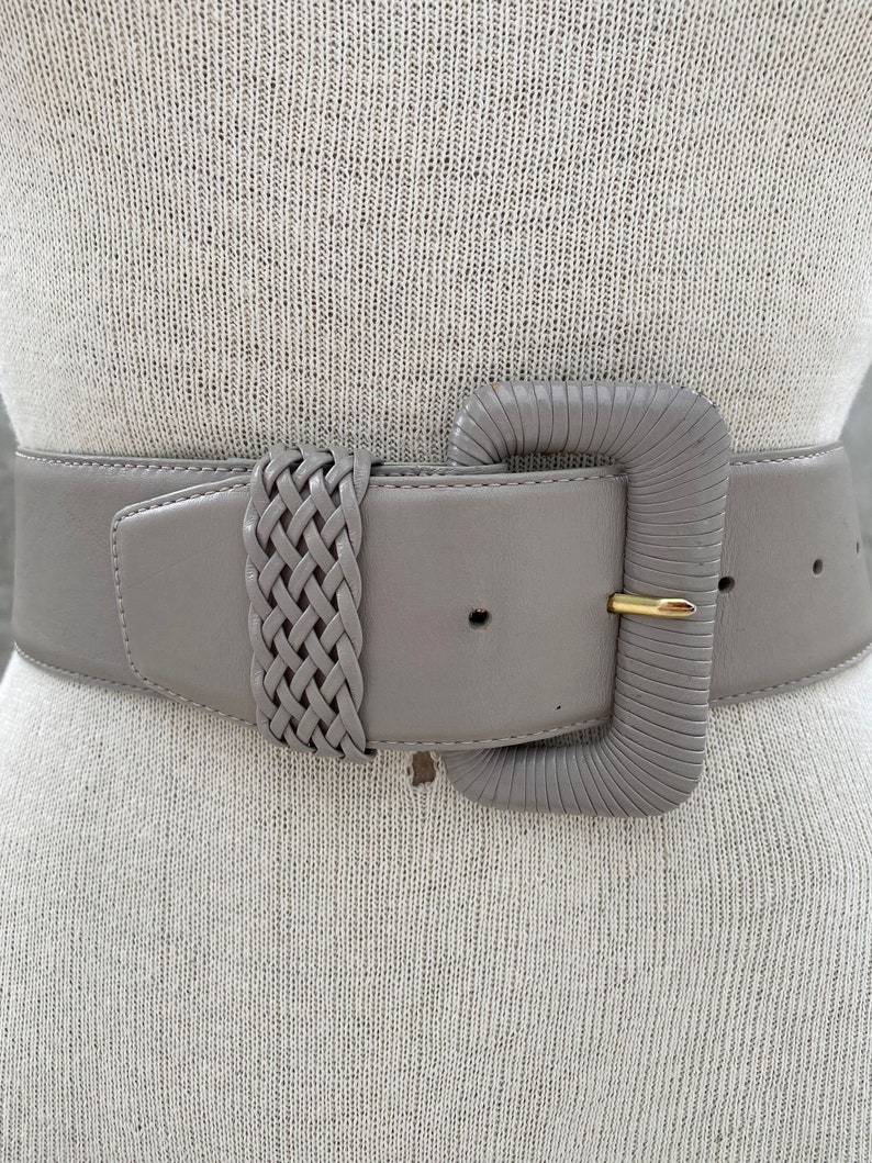Puede incluir: Un cintur&oacute;n ancho de cuero gris con un detalle trenzado y una hebilla rectangular grande de color gris. La hebilla tiene un pasador de metal dorado.