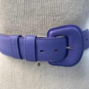 May include: A close-up of a purple belt with a textured surface. The belt has a large, square buckle and a matching belt loop. The belt is made of a durable material and is designed to be worn around the waist.