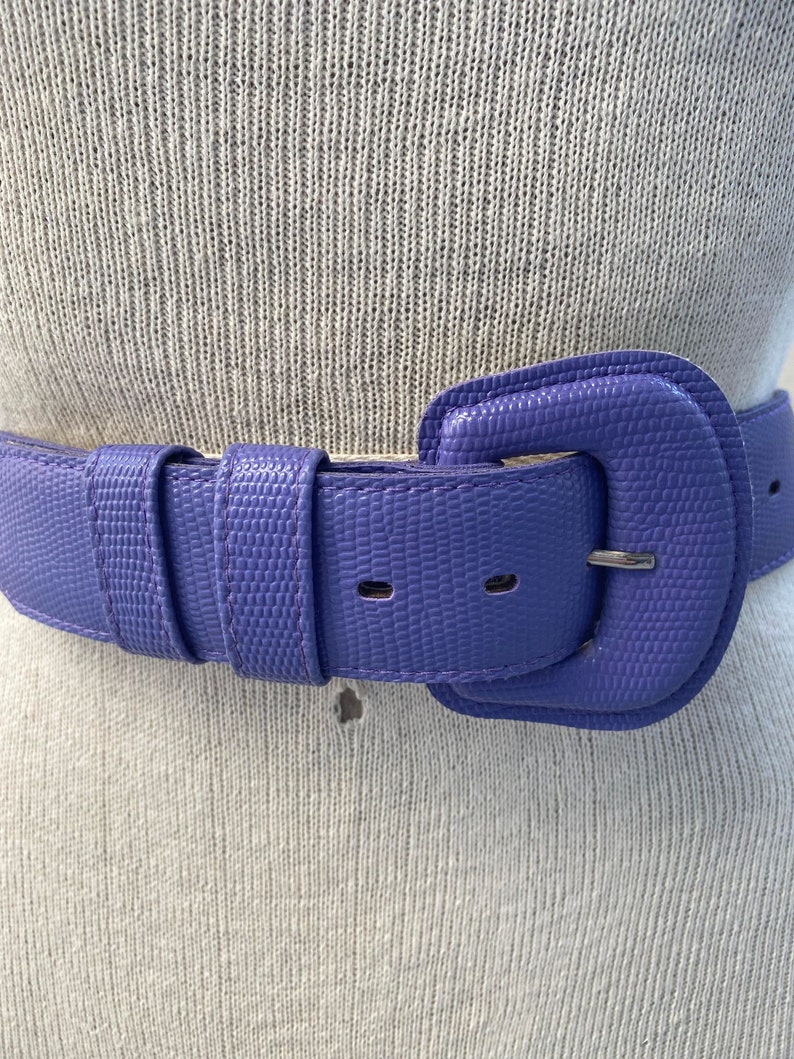 May include: A close-up of a purple belt with a textured surface. The belt has a large, square buckle and a matching belt loop. The belt is made of a durable material and is designed to be worn around the waist.