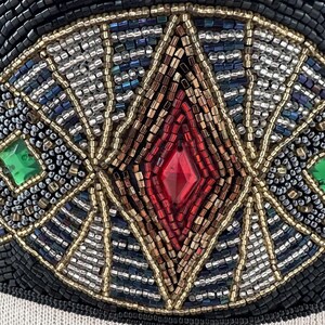 Pu&ograve; includere: Primo piano di un disegno di perline con un gioiello rosso a forma di diamante circondato da perline dorate, argentate e blu. Il disegno &egrave; su uno sfondo nero.