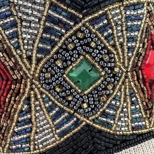 Pu&ograve; includere: Primo piano di un disegno a perline con una gemma verde a forma di diamante al centro. Il disegno &egrave; composto da piccole perline colorate in un motivo geometrico.