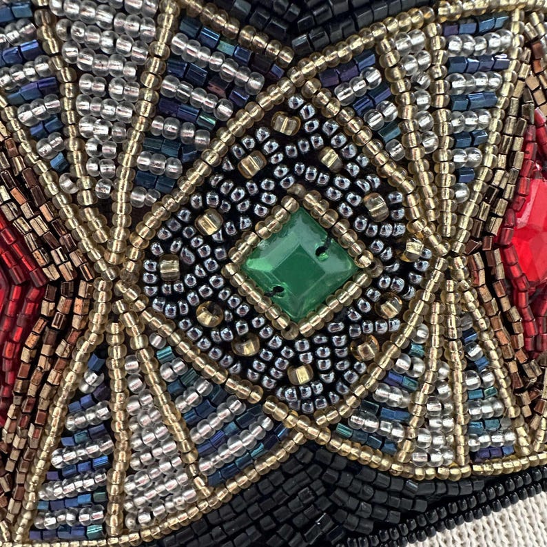 Pu&ograve; includere: Primo piano di un disegno a perline con una gemma verde a forma di diamante al centro. Il disegno &egrave; composto da piccole perline colorate in un motivo geometrico.