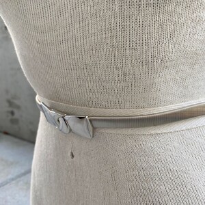 May include: A silver metal belt with a decorative buckle. The belt is on a white knitted fabric.