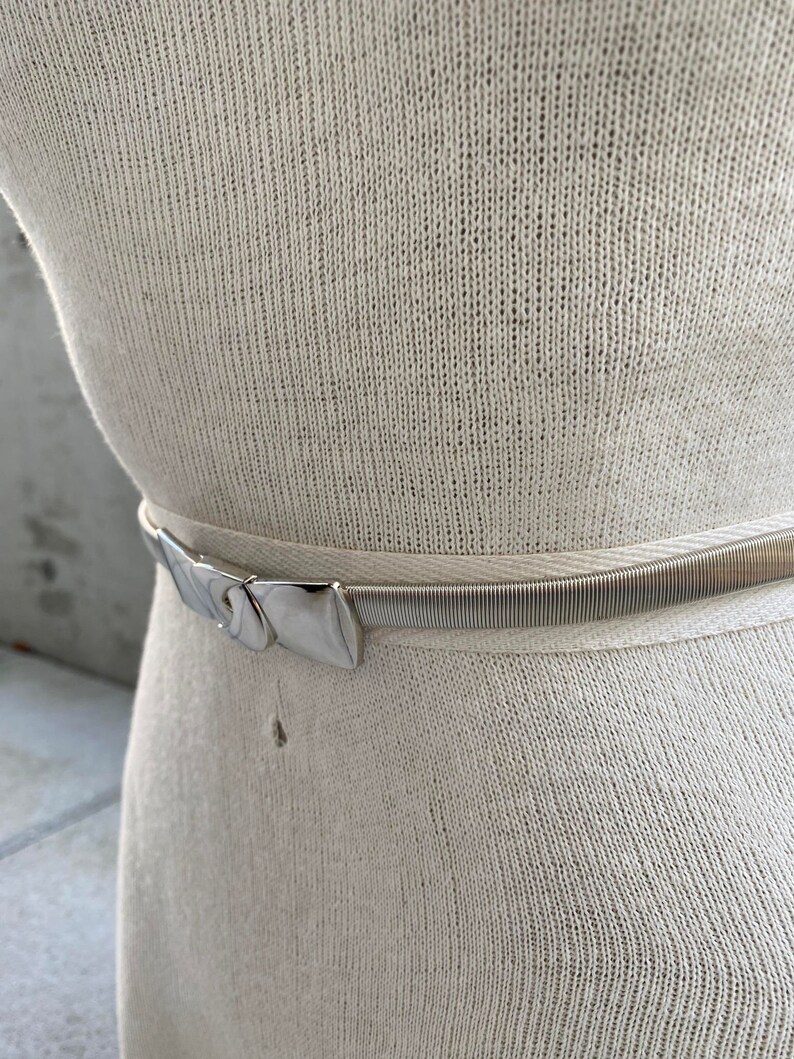 May include: A silver metal belt with a decorative buckle. The belt is on a white knitted fabric.