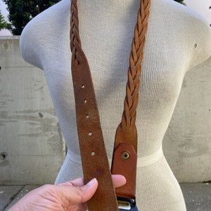 May include: A brown leather braided belt with a metal buckle. The buckle has a design on it.