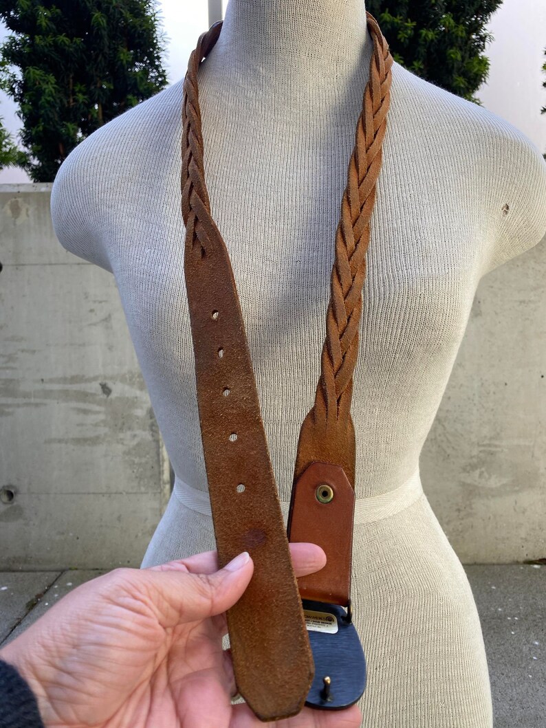 May include: A brown leather braided belt with a metal buckle. The buckle has a design on it.